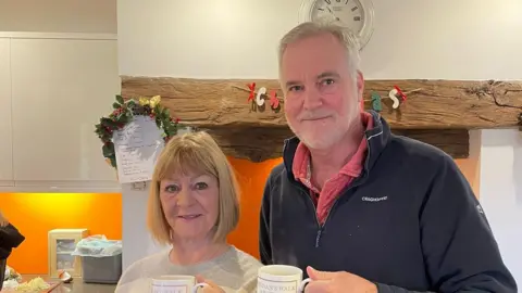 Brendan Wilson A cosy kitchen scene, Celia and Brendan are standing side by side, holding matching white mugs with printed text on them. They are positioned in front of a large wooden beam and an inglenook‑style fireplace adorned with a small festive wreath and decorative letters spelling &ldquo;Christmas.&rdquo; Behind them are clean white cabinets and a lit orange wall. The atmosphere is warm and homely.