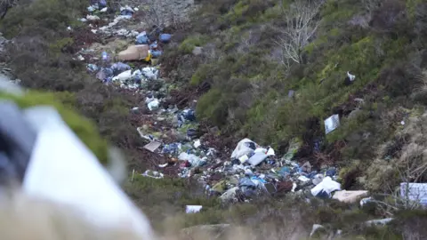 Nathan Dixon Fly-tipped waste on the side of a green mountain. There are shrubs either side, with the waste piled in a ditch running down the slope