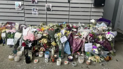 Many bunches of flowers, with candles in the foreground. Photographs are stick on a slatted wall which have been left in tribute to the two men.