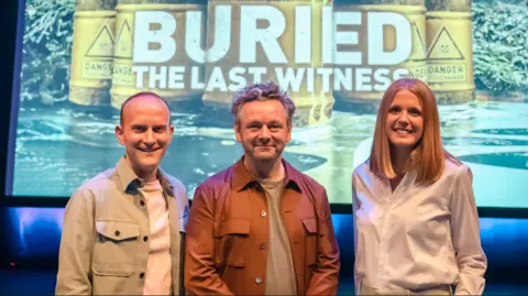 The podcast logo is on the screen in the background. Sheen stands in the centre. He is wearing a brown t-shirt and a copper jacket. Ashby on the left wears a light green jacket over a white t-shirt and Taylor wears a white shirt.