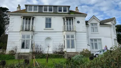 Talland House viewed from the front. It looks like a French-style villa with a more recent extension to the right. There are three storeys. There are bay doors on the ground floor and the windows on the first floor have balconies. The house is white. The garden extends in front of it.
