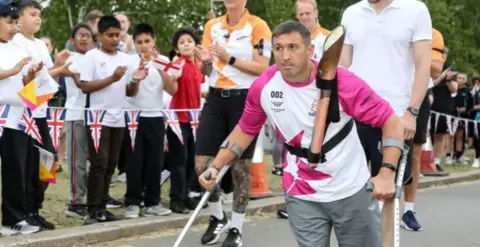 Getty Images Ben Parkinson during the Queen's Baton relay in Birmingham 2022 to mark the Commonwealth Game - he is wearing a pink and white t-shirt and using crutches with a determined look on his face. Visible behind him are a crowd of children cheering him on standing behind a cordon made up of miniature union flags