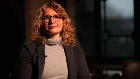 Dr Kelly Johnson is sitting in a room. She has long curly hair and is wearing a suit jacket over a roll neck jacket and has glasses.