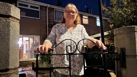 Kevin Shoesmith/BBC A woman in her mid 70s, with thinning brown hair, poses for the camera in front of a black metal gate