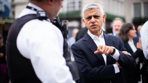 PA Media Sir Sadiq Khan, in a dark blue suit with a white open necked shirt, stands in the street talking to a police officer.