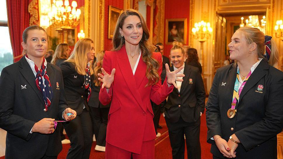 The Princess of Wales is wearing a red suit and holding her arms up as she speaks with members of England's Women's World Cup-winning team