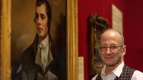 Nick Mailer Dr Bill Zachs, who is bald and wearing a red, yellow and dark checked waistcoat, a white shirt and mustard-coloured glasses, stands next to the re-discovered portrait of Robert Burns by Henry Raeburn which is hanging in the National Galleries of Scotland
