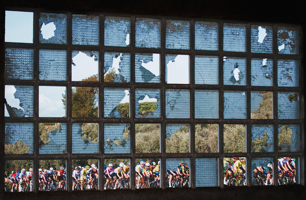 A large group of cyclists in colorful jerseys seen through a shattered glass window. The broken panes frame the riders against a backdrop of trees and sky.
