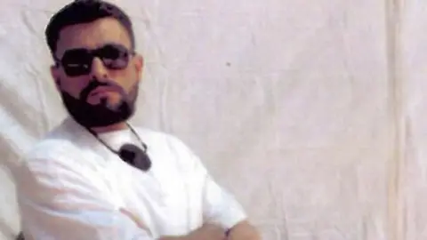 Abu Zubaydah wearing a white t-shirt and white trousers, carrying an eye-patch around his neck. He is also wearing black sunglasses and has a defined beard.