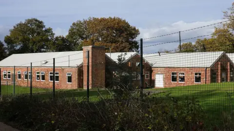 PA Media The barracks are seen behind a barbed fence - a group of buildings set over one floor made of brick with white rooves, taken in October 2025.