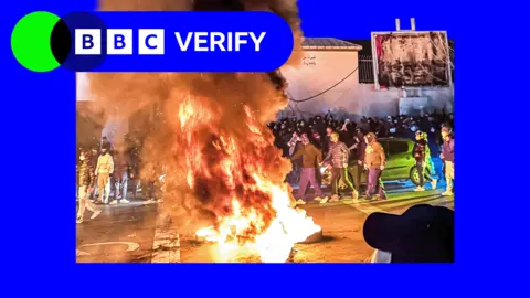 BBC Huge fire and plume of smoke in the centre of the image with crowds of masked people surrounding it during Tehran protest