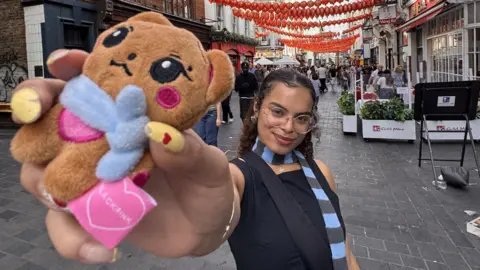 Ashia Davies Ashia is wearing a black top with a black and blue scarf. She is holding  a small teddy up to the camera. She has dark curly hair and brown eyes and glasses.