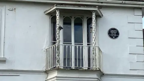 The exterior of a set of French windows with a balcony. A black plaque commemorating Virginia Woolf is mounted on the wall to the right of it.
