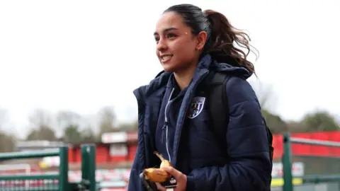 Getty Images Mariam Mahmood is seen at a football training ground wearing a blue waterproof jacket and carrying a backpack. She has long dark hair tied up. In the background there is red seating in the stands and a green fence.