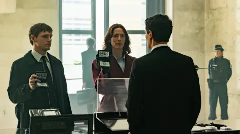 Disney via Getty Images Evan Peters' character wears a shirt, tie and jacket and is holding up his FBI ID card and Rebecca Hall's character wears a red jacket, shirt and also holds up an FBI ID card in front of a man, whom we only see the back of.