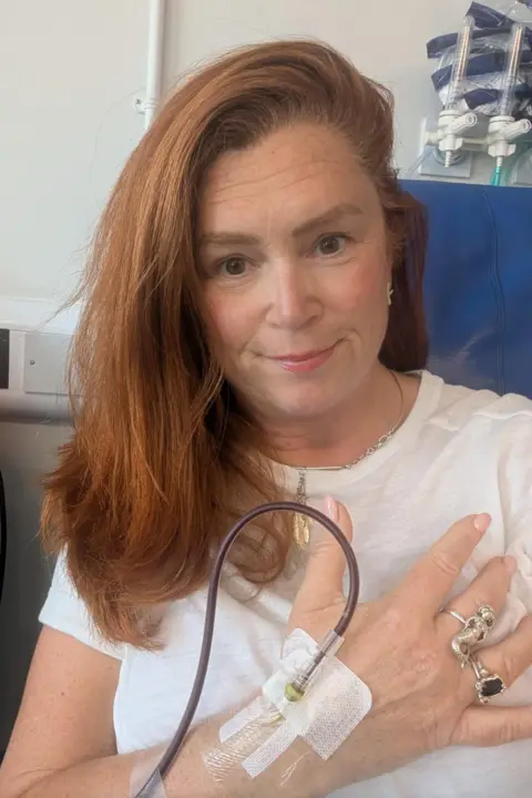 Sian Chathyoka Selfie of Sian sat in a blue hospital chair with a drip going into her hand. She is wearing a white t-shirt and has straight ginger hair.