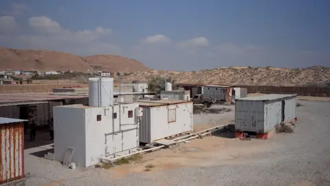 Liam weir / BBC One white shipping container and two grey ones are seen on a gravel area with sandy hills behind them. A small white building with a water tank on it is next to one of them.