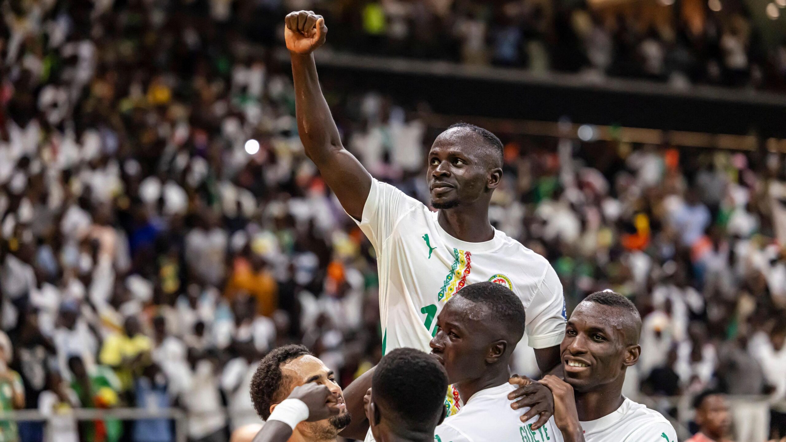 Sadio Mane holds his right fist aloft in celebration as he is held aloft by Senegal team-mates after scoring a goal. Behind him in the distance a large crowd can be seen out of focus in the stands