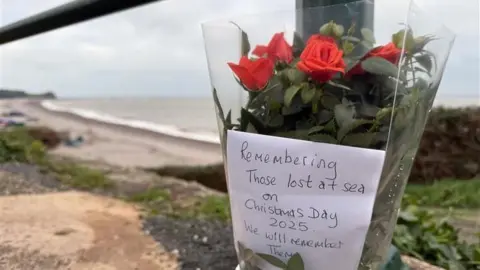 A floral tribute tied to a pole. On the distance there is a beach. The flowers are red and there is a note in the flowers with the words 'Remembering those lost at sea on Christmas Day 2025. We will remember them.'