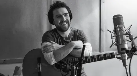 Danielle MacLeod Michael McGovern in the studio, with a guitar resting on his lap and a  microphone set up in front of him. He has dark hair,  a beard and a goatee.