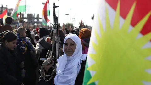 AFP A woman holds a rifle at a protest by Kurds against a government offensive in north-eastern Syria, in Qamishli, Syria (20 January 2026