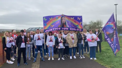 A group of family and friends wearing 'justice for Chloe' t-shirts and banners. They are standing in a park.