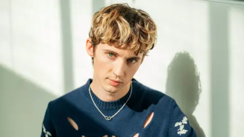 BBC/Sarah Louise Bennett A portrait of Troye Sivan where he is looking at the camera with brownish hair. He has a nose ring on, a blue jumper and a necklace. He's infront of a white wall.