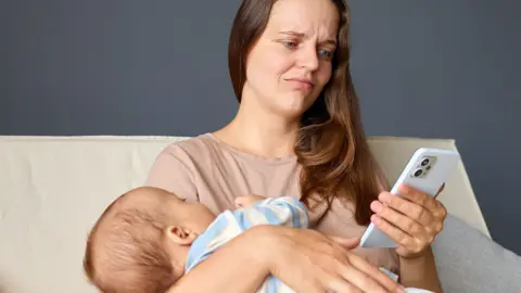 SementsovaLesia/GettyImages A woman sitting on a cream sofa holding a baby dressed in striped clothing. The person is using a smartphone with their free hand while supporting the baby in their lap. A dark gray wall is visible in the background.