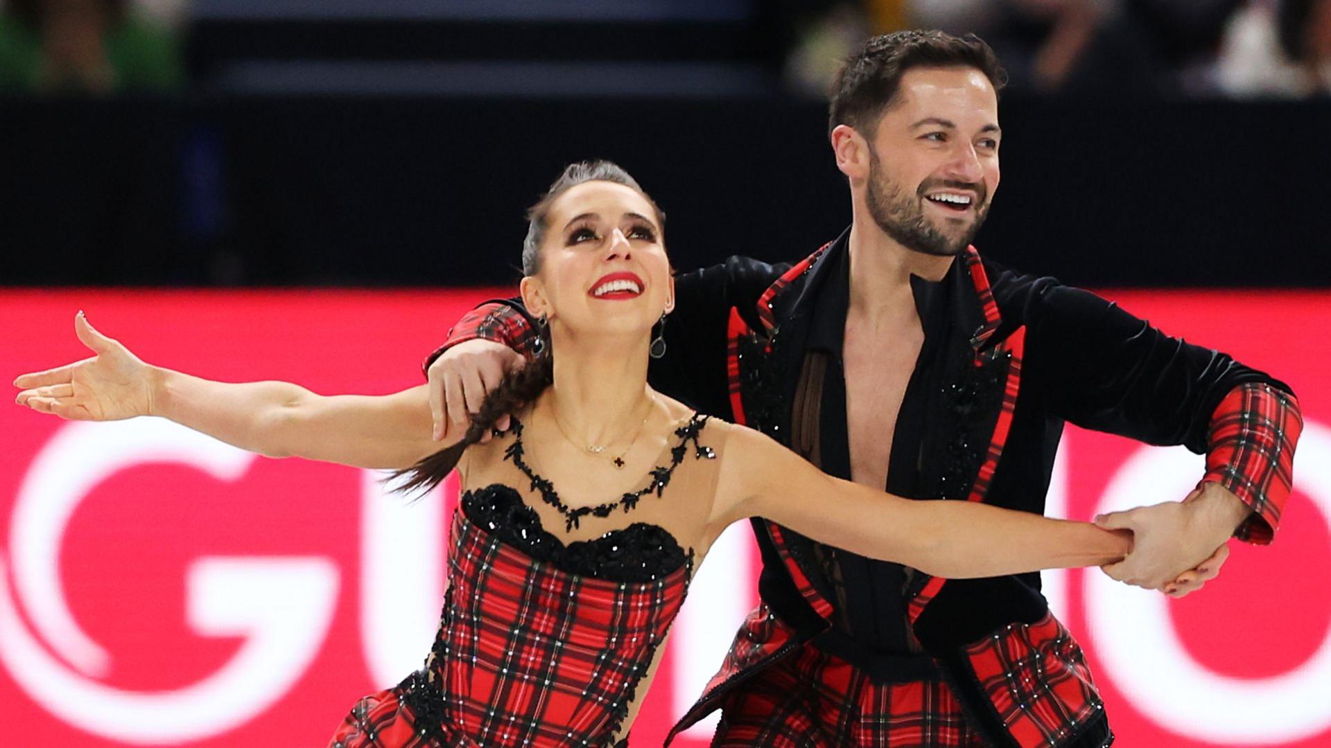 Figure skating pair Lilah Fear and Lewis Gibson perform a routine together, with both dressing in black and tartan outfits, with both smiling