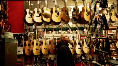 A wall has more than a dozen classic guitars hanging from it in two lines. A man with a beard and short hair, in a black coat, is in the bottom of the frame, looks up to the right.