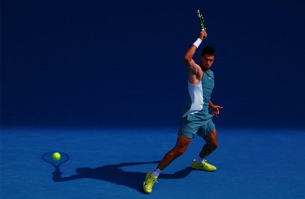 Tennis player Carlos Alcaraz hits the ball during his fourth round match against Britain's Jack Draper. He is wearing a teal outfit and neon yellow shoes on a bright blue court, with his shadow appearing as though it is hitting the ball.