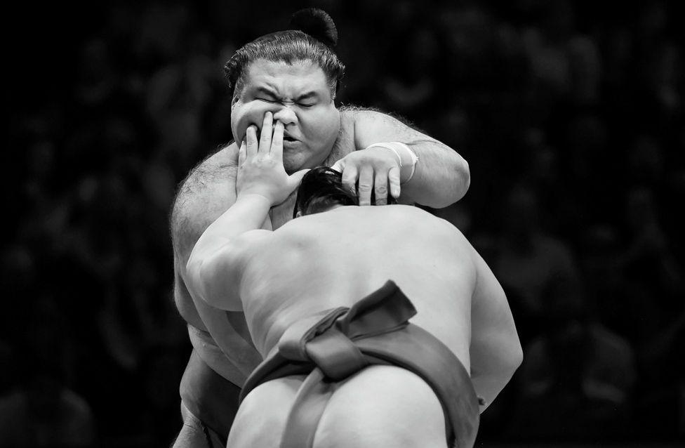 Two sumo wrestlers grapple in a traditional ring, captured in black and white. One wrestler pushes forward while the other braces, with the crowd blurred in the background.