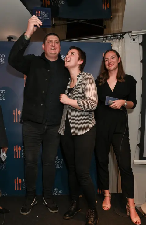 Getty Images Chef Gareth Ward stands on a small stage in front of a &ldquo;The Good Food Guide&rdquo; backdrop. He raises an award plaque above his head while smiling, his guest, a woman, looks up at it, smiling, and a woman on the right smiles while holding a microphone and a card.