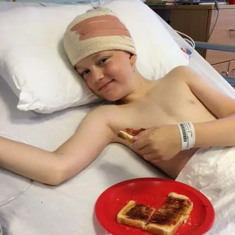 Claire Lindsay A young boy in a hospital bed with his head wrapped in bandages. He has a hospital ID bracelet around his wrist and his head is resting on a white pillow. He has a red plate and some toast on the bed next to him. He has a piece of toast in his hands.