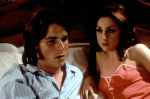 Alamy Christian Bale, dark hair and side burns, as Chris in bed in striped top with Elsa Zylberstein, who played his French girlfriend Annick, in strappy orangy red neglige