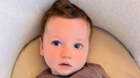 Dave and Siobhan Smith A baby boy with big blue eyes is looking just away from the camera. He has a brown top and is lying on a support cushion.