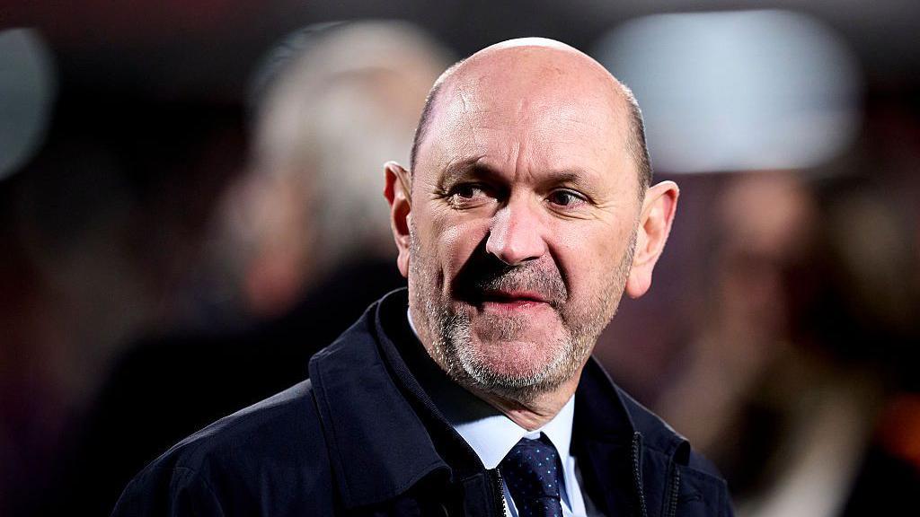 A close-up image of Rafael Louzan. He is bald and has a greying, stubbly beard. He is wearing a navy tie with a light blue shirt and dark coat.