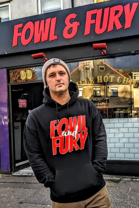 Fowl and Fury Jamie Rees standing in front of Fowl and Fury in Cathays. The red lettering on the black building. Jamie is wearing a beanie, black hoodie. Fowl and fury lettering on the hoodie.