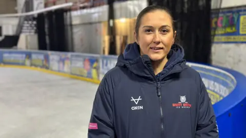 A woman in a thick jacket with a Great British Ice Hockey logo on the left side stands in front of an ice rink. The surface is marked with tracks from multiple skates.