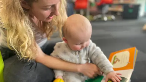 A woman with wavy blonde hair sits on the floor with an arm around her baby, who is touching a book.