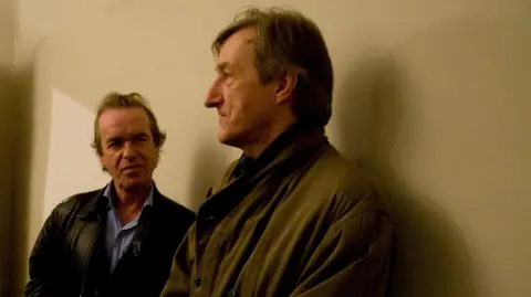Dafydd Jones Martin Amis on the left  -- in a black leather jacket and pale blue shirt - looking at Barnes -- in profile -- in khaki jacket. Both standing against a beige wall