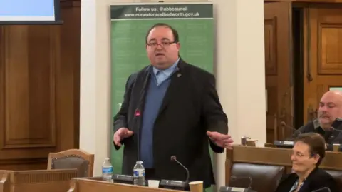 Kris Wilson stands in the council chamber and addresses the meeting. He is wearing a dark suit jacket and light blue jumper and shirt.