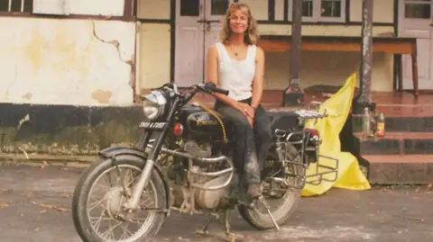 Jacqui Furneaux Jacqui Furneaux sitting on her motorbike smiling outside a rundown house on her travels in India