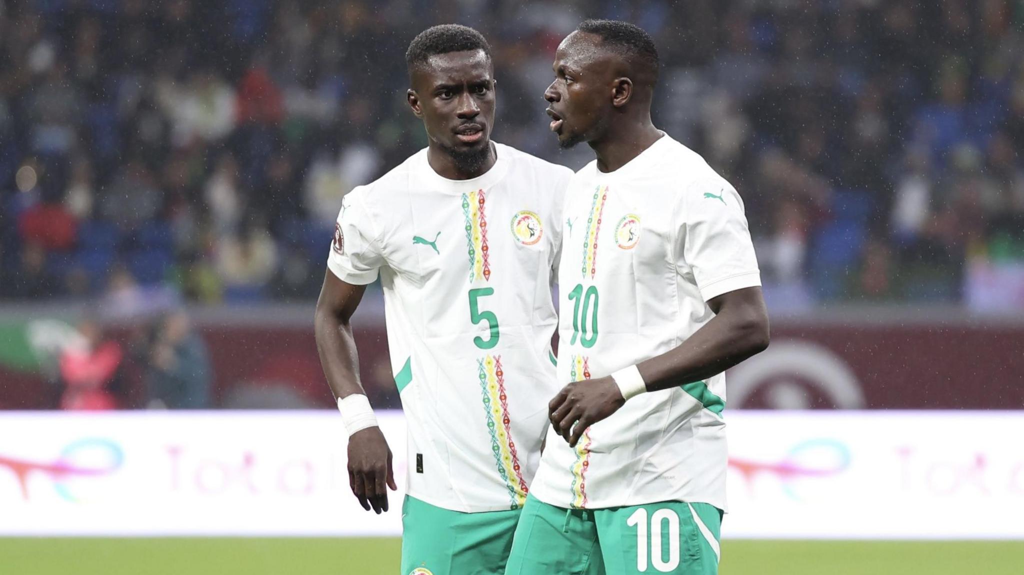 Idrissa Gana Gueye stands talking to Sadio Mane during a football match. Both players wear white shirts bearing green, yellow and red detail on the front and green shorts. Gana Gueye's shirt has the the number five on the chest while Mane's kit bears a 10 on the chest and left short leg