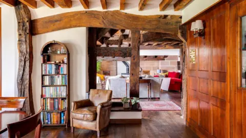Matt Elson A reading nook within a historic home featuring exposed dark timber beams and wooden floors. A tall narrow bookshelf stands in an arched alcove filled with colourful books and small ornaments. A brown leather armchair sits nearby. Through an open timber-framed partition, another room is visible with sofas, rugs, and more beams, creating a warm and inviting interior.