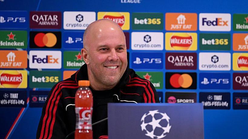 Arne Slot, in a black top with three red stripes on each arm, talks to the media a day before Liverpool's match against Qarabag, with a wall of sponsor logos behind him