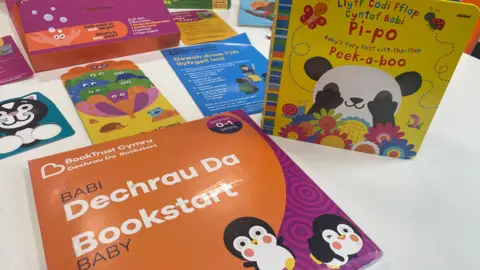 Baby books laid out on a white table, including Bookstart Baby which is orange and purple with pictures of penguins in the foreground, a peekaboo book and some information leaflets about accessing your local library and the Bookstart scheme.