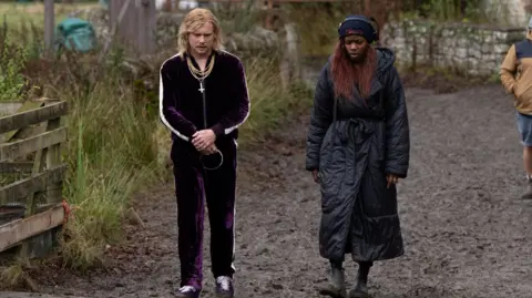 Sony Images Jack O'Connell and Nia DaCosta on the set of 28 Years Later: The Bone Temple - he wears a purple velour tracksuit and gold chains and has shoulder length blond hair, she has a long black padded coat and short boots - they are looking down and walking outside on a muddy path