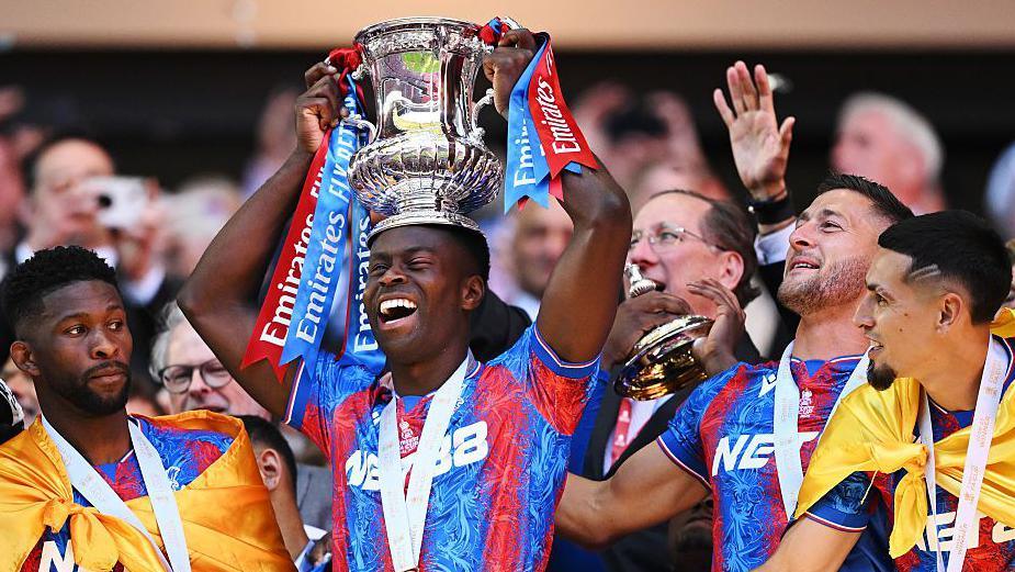 Guehi lifts the FA Cup trophy onto his head as teammates watch on at Wembley
