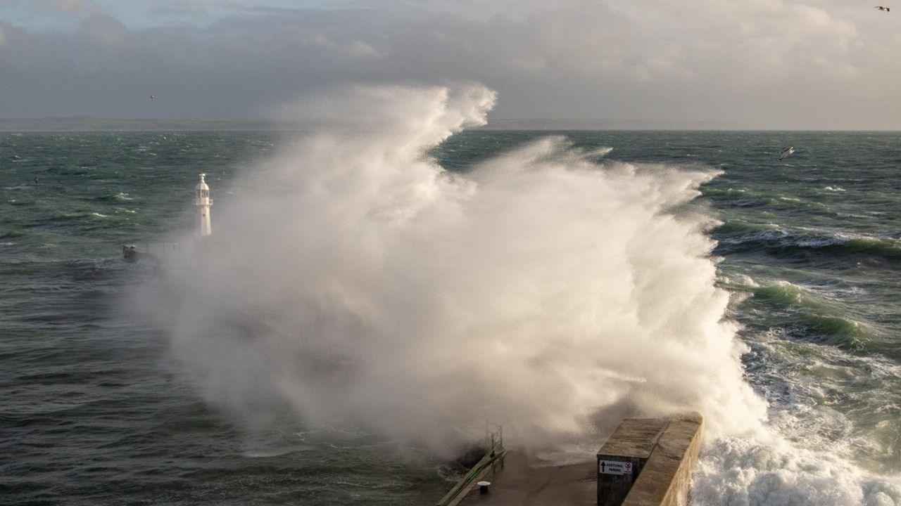 A large wave crashes against a breakwater with a lighthouse sending up a big spray of foam